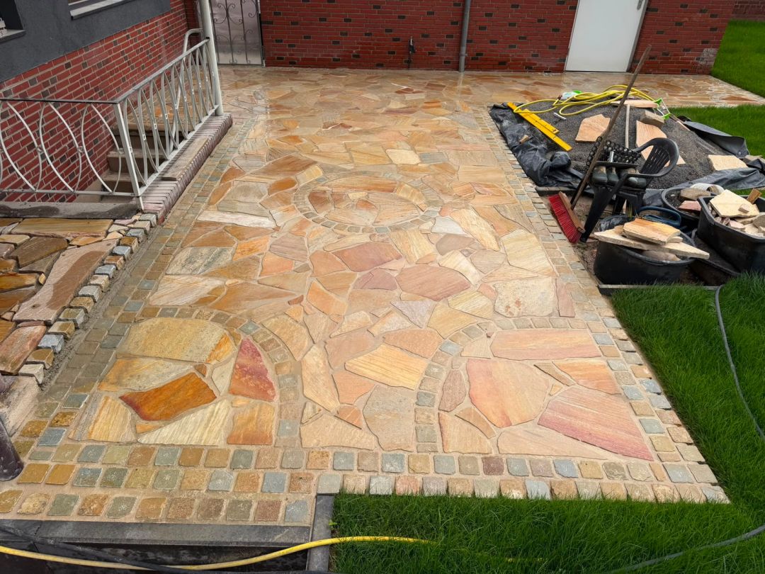 Neu verlegte Natursteinterrasse mit rundem Mosaikbogen und gepflastertem Rand an einer Hauswand