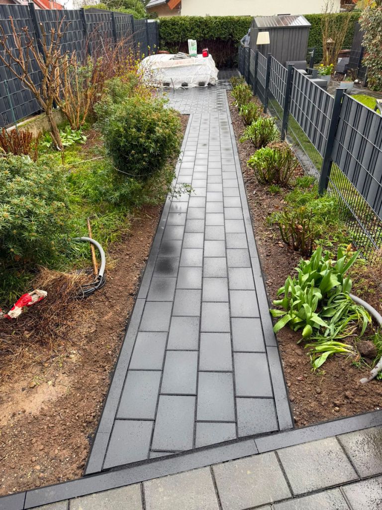 Schmaler Gartenweg aus grauem Betonsteinpflaster mit Randeinfassung zwischen zwei Beeten