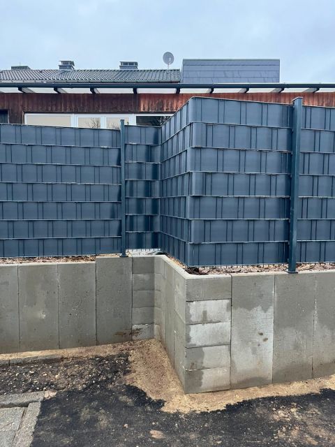 Über Eck geführter Doppelstabmattenzaun mit Sichtschutz auf einer Mauer aus Betonsteinen, davor ein noch offener Bodenstreifen