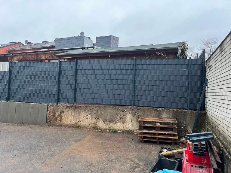 Lange Zaunanlage mit Sichtschutzstreifen auf einer Betonsockelmauer entlang eines Hofes, rechts eine gemauerte Wand